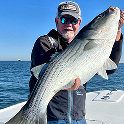 A man on a boat, wearing a ball cap and sunglasses, holding a large striped bass